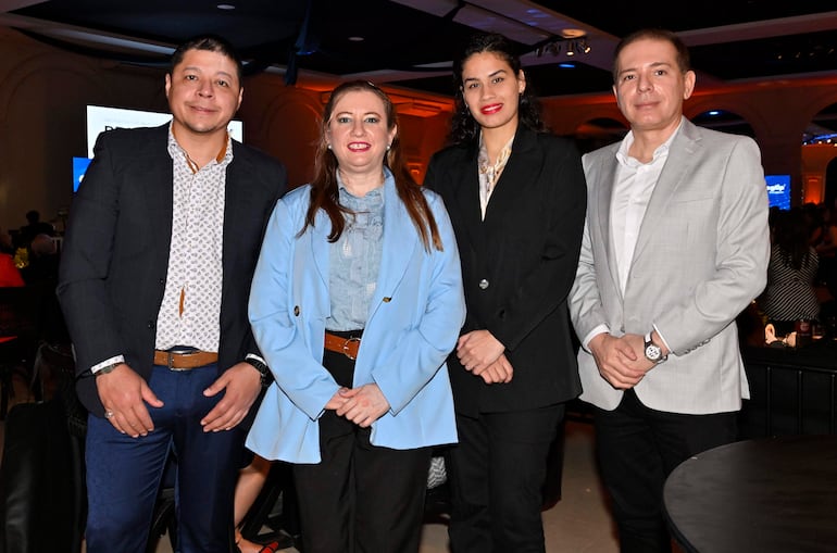 Cuatro personas sonríen, con la mujer del blazer azul al centro, en un salón bien iluminado durante un evento social.