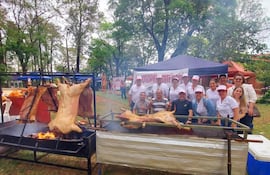 Grupo de personas con camisetas blancas y gorras sonríe frente a una parrilla con asados en un parque arbolado.