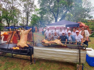 Grupo de personas con camisetas blancas y gorras sonríe frente a una parrilla con asados en un parque arbolado.