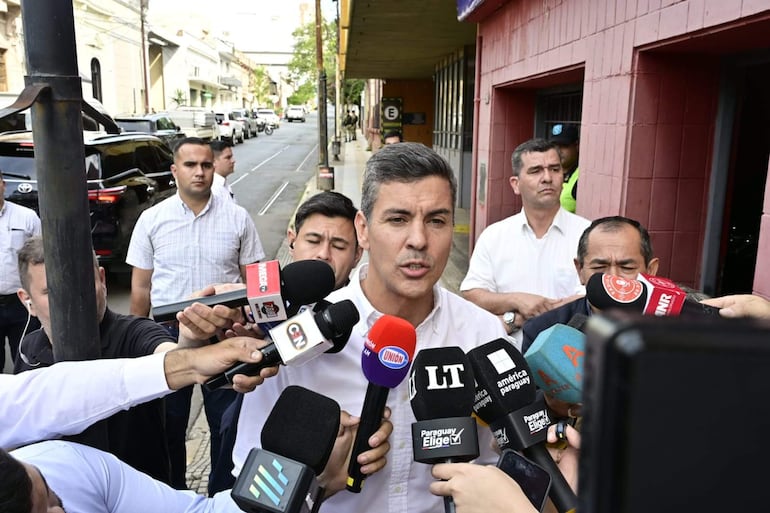 Santiago Peña con micrófono, rodeado de periodistas en una escena urbana, reflejando emociones de tristeza y tranquilidad.