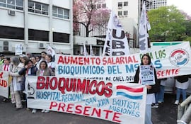 Un grupo de bioquímicos en ropa blanca sostiene pancartas en una protesta, destacando a una mujer con un cartel sobre el Hospital Regional San Juan Bautista.