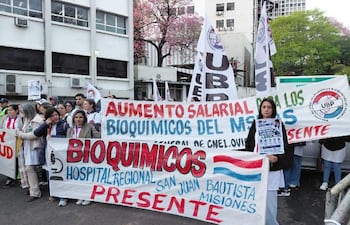 Un grupo de bioquímicos en ropa blanca sostiene pancartas en una protesta, destacando a una mujer con un cartel sobre el Hospital Regional San Juan Bautista.