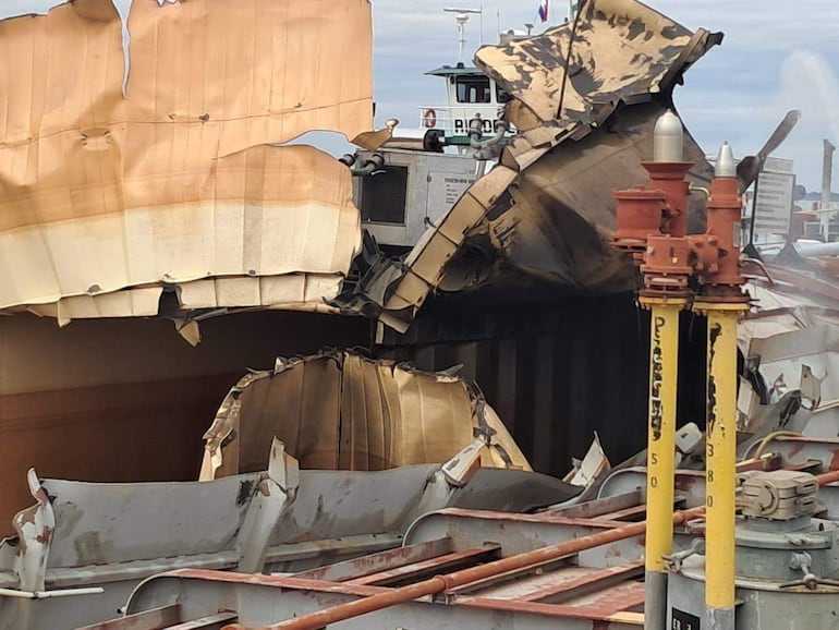 Barco con daño visible en cascarón, amarrado en puerto, sin personas presentes, entorno marítimo nublado.