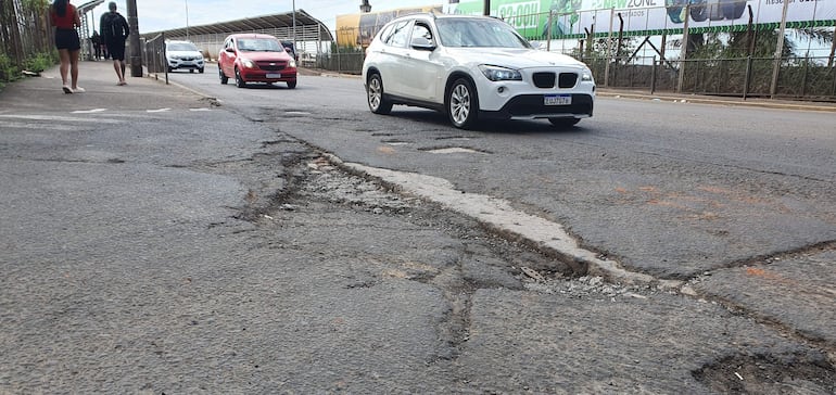 Dos peatones, uno con camiseta negra y otro con mochila, caminan junto a un bache en la carretera, con coches circulando.