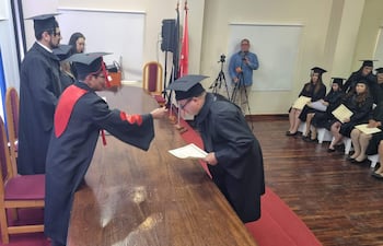 Graviel Benítez de pie con birrete y toga, recibe la borla de un oficial en ceremonia con fondo decorado en colores institucionales.