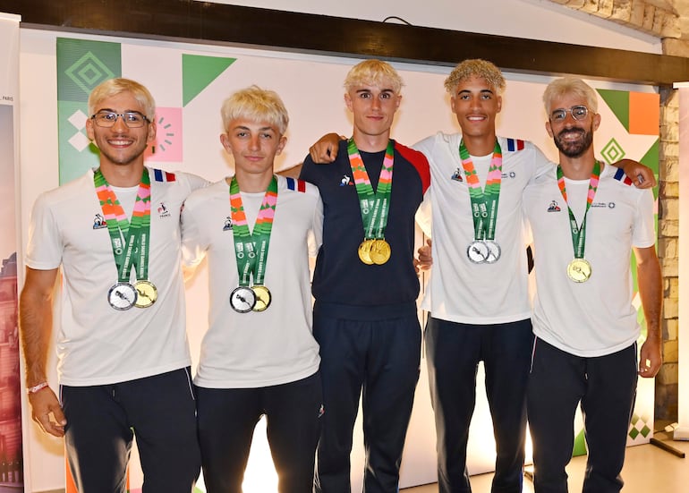 Cinco hombres con camisetas blancas y medallas en un evento festivo; expresiones felices y ambiente decorado.