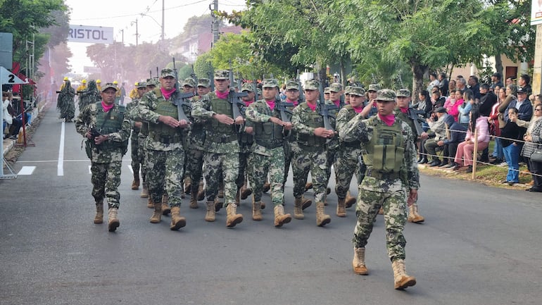 Grupo de 15 militares en formación, vestidos con uniformes camuflados y pañuelos rosas, marchando por la calle en un desfile.