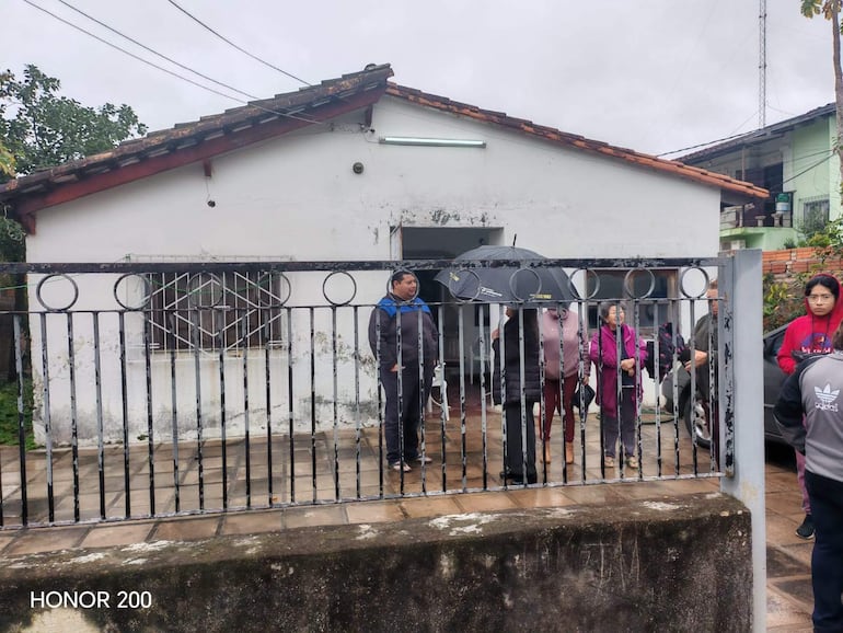 Grupo de cinco personas esperando en el exterior, una mujer con paraguas negro, joven en sudadera roja, clima nublado.