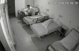 Cuidador vestido de blanco en clínica, ajustando la ropa de un paciente en una cama vacía del hospital.