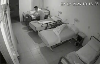 Cuidador vestido de blanco en clínica, ajustando la ropa de un paciente en una cama vacía del hospital.