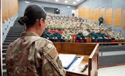 Mujer con uniforme militar de espaldas en atril, hablando ante audiencia de hombres en uniformes de camuflaje, en ambiente formal iluminado.