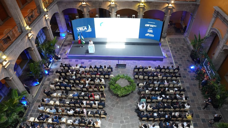 Gran salón lleno de personas sentadas, mientras un orador habla en el podio con logotipos del evento y banderas.