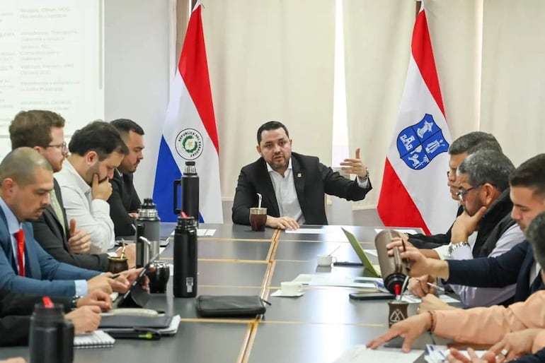 Luis Bello con chaqueta negra y camisa blanca gesticula en reunión, rodeado de hombres en una oficina decorada con banderas de Paraguay.