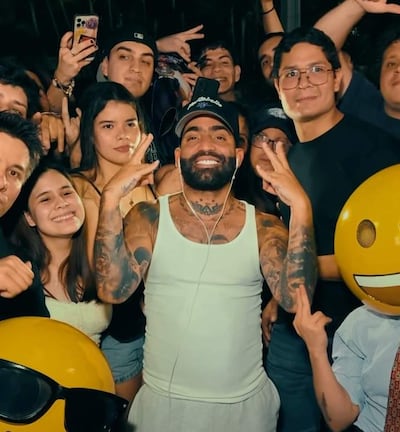 Arcángel, con camiseta sin mangas y gorra, sonríe con fans que hacen señales de paz en un ambiente festivo.