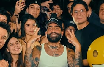 Arcángel, con camiseta sin mangas y gorra, sonríe con fans que hacen señales de paz en un ambiente festivo.