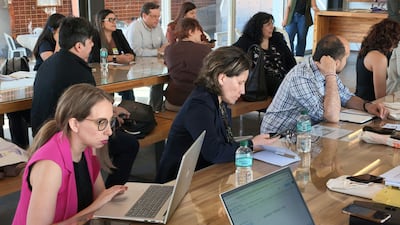 Mujeres en blusa negra y chaleco rosa, con laptop y teléfono, rodeadas de documentos sobre la mesa en ambiente de reunión.