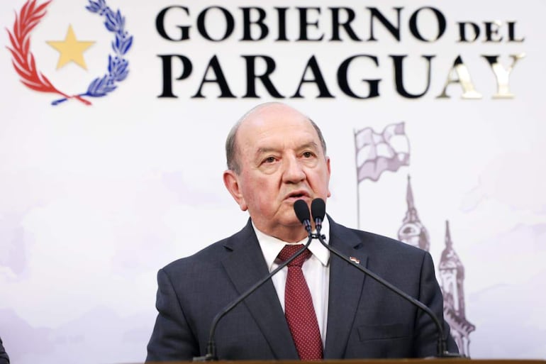 Hombre en traje oscuro con corbata de puntos rojos, hablando en un acto oficial con fondo de 'Gobierno del Paraguay'.