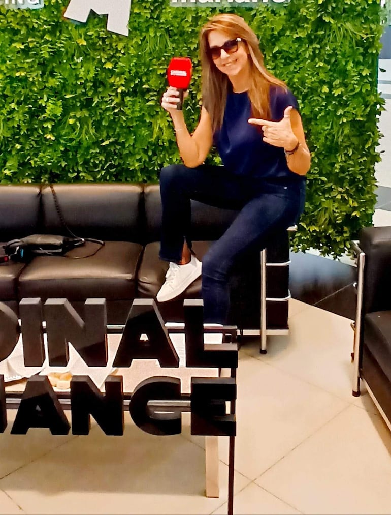 Mujer con top oscuro y jeans, gafas de sol, sonriendo y sosteniendo un micrófono rojo en un ambiente decorativo.