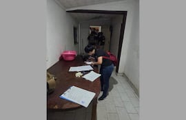 Mujer con camiseta negra y mochila roja revisa documentos sobre una mesa en una habitación iluminada tenue.