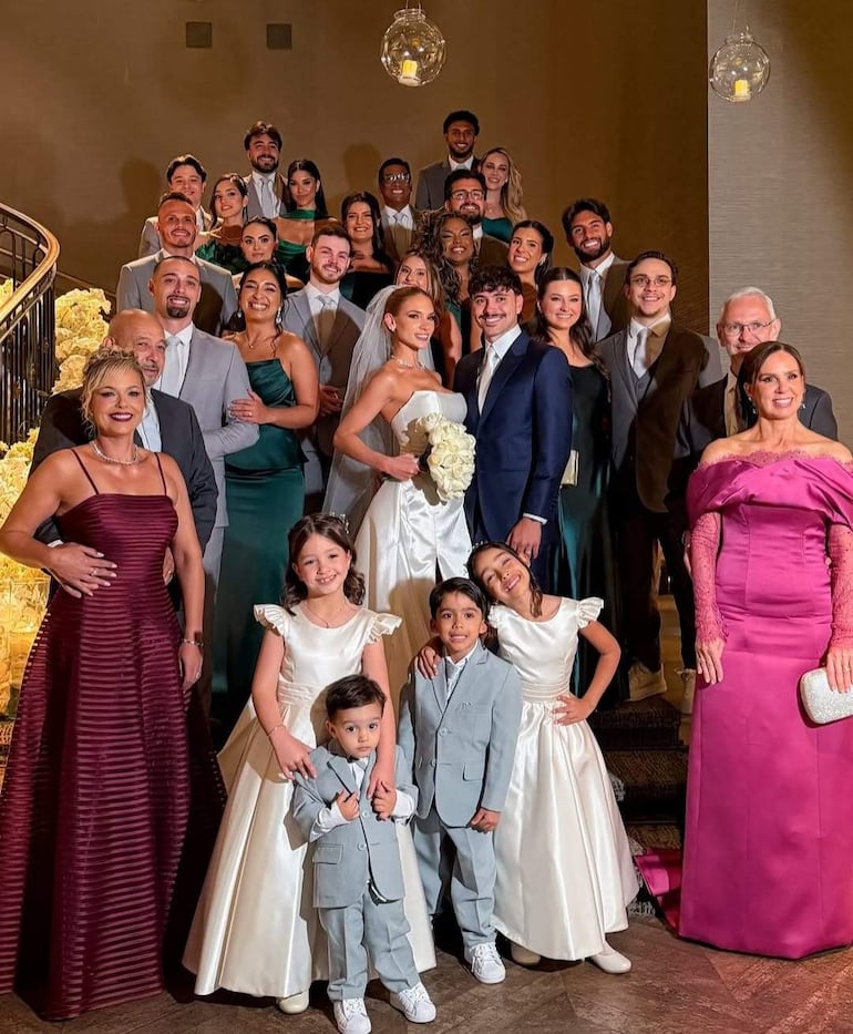 Novia en vestido blanco con ramo, rodeada de invitados elegantes en la escalera, todos sonrientes y felices.