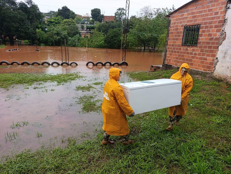Dos hombres con chaquetas impermeables amarillas transportan un equipo en un entorno inundado con agua y hierba.