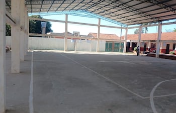 Cancha deportiva techada y vacía con suelo de cemento, iluminada por luz natural y portería de fútbol al fondo.