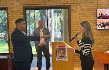 Hombre de traje oscuro jurando, junto a mujer con micrófono y documento, en un ambiente iluminado con ventanas y vegetación de fondo.