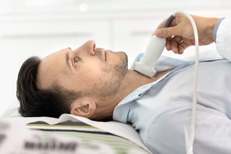 Hombre en mesa de examen, mientras un profesional le aplica ultrasonido en el cuello.
