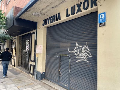Persona con abrigo oscuro y pantalones claros camina frente a la joyería Luxor con persiana cerrada y grafitis visibles.