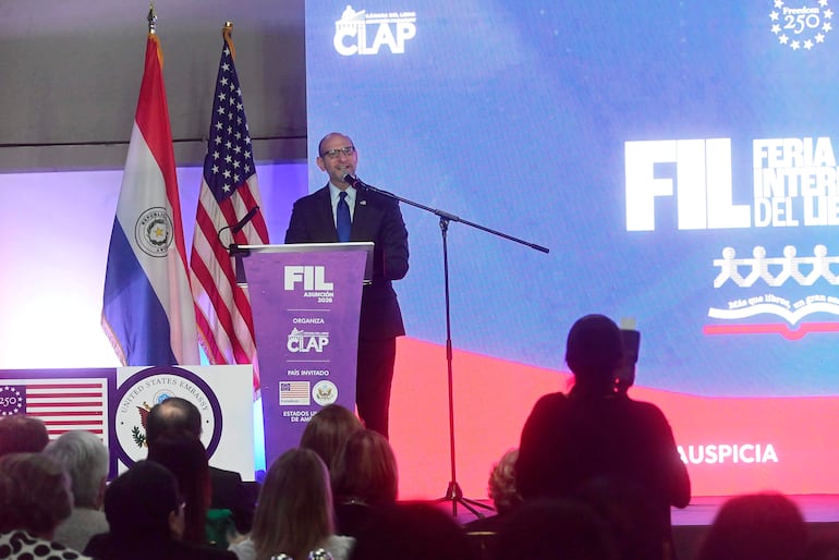 Hombre en traje oscuro frente a un podio con banner de FIL Asunción 2026, banderas de Paraguay y Estados Unidos en el fondo.