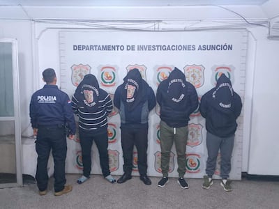 Cinco hombres, cuatro encapuchados y un policía supervisando, en un interior identificable con logo del Departamento de Investigaciones Asunción.