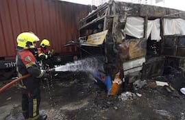 Bomberos tratan de extinguir el fuego en los restos del bus impactado por un tren, este sábado en Bangkok.