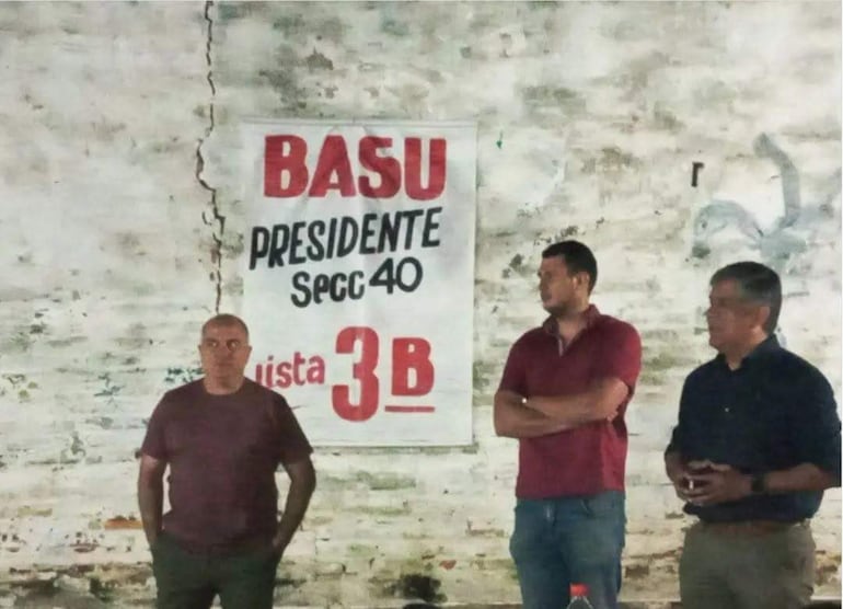 Tres hombres en camiseta negra, roja y camisa oscura conversan en un ambiente simple con pancarta de 'BASU PRESIDENTE'.