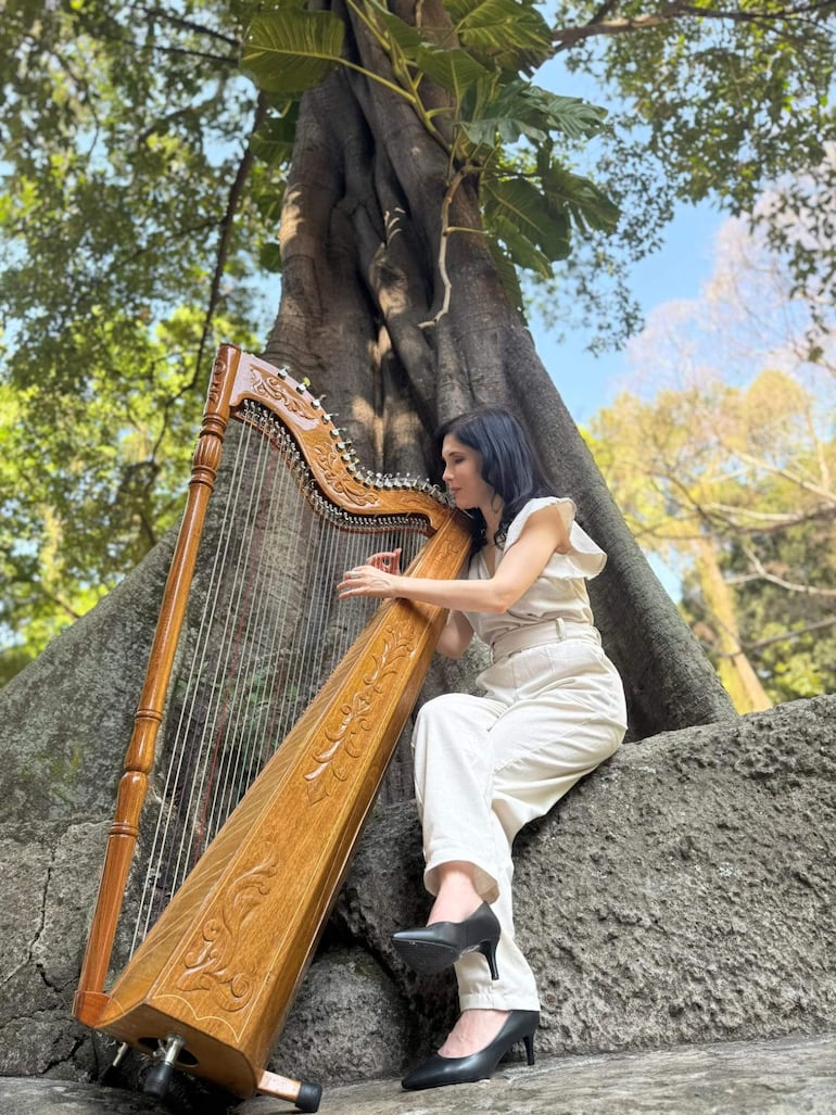 Mujer con cabello oscuro y lacio, vestida con un conjunto claro, tocando un arpa en un entorno natural.