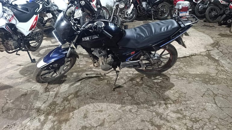 Motocicleta Kenton negra y azul estacionada en un parqueadero, sobre su pata de cabra, sin presencia de personas.