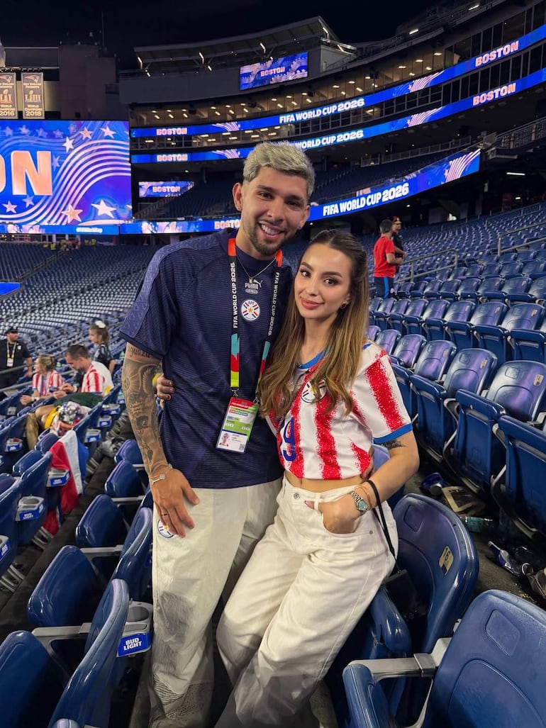 Ramón Sosa con camiseta oscura y gafete, y Julio Enciso con top a rayas, sonriendo en un estadio lleno de aficionados.
