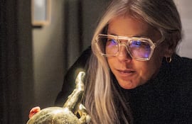 Laura Mandelik, con cabello rubio y gafas grandes, inclina su cuerpo hacia un objeto dorado iluminado en un entorno oscuro.
