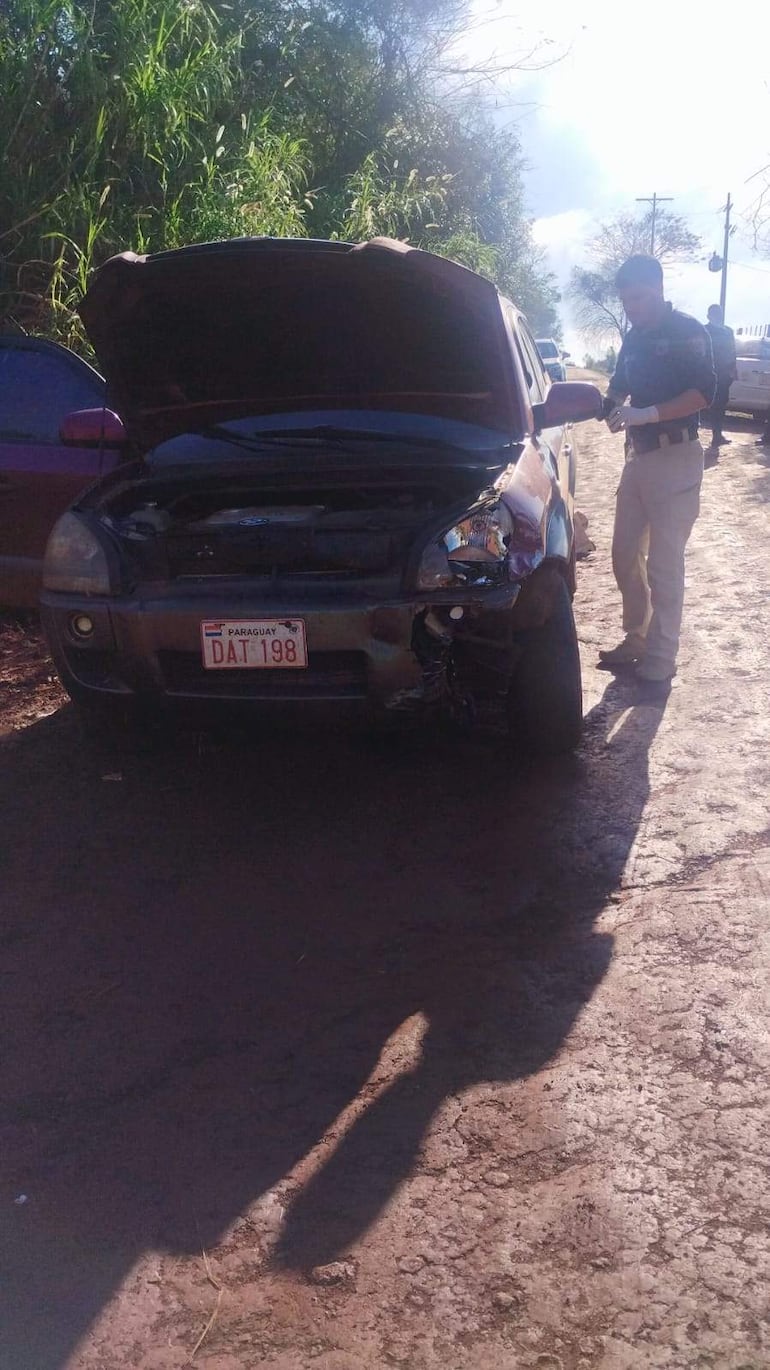 Policía cerca de un vehículo averiado en camino de tierra, con capó levantado y daños visibles en la parte frontal derecha.