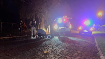 Escena con luces de ambulancia iluminando un camino, cuatro personas presentes, una en uniforme camuflado, otra agachada en el suelo.