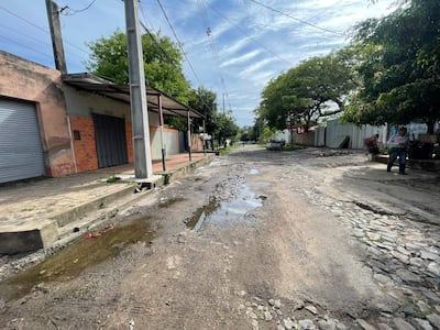 Una persona con camisa clara y jeans camina mientras la calle de tierra y piedras muestra charcos de agua.