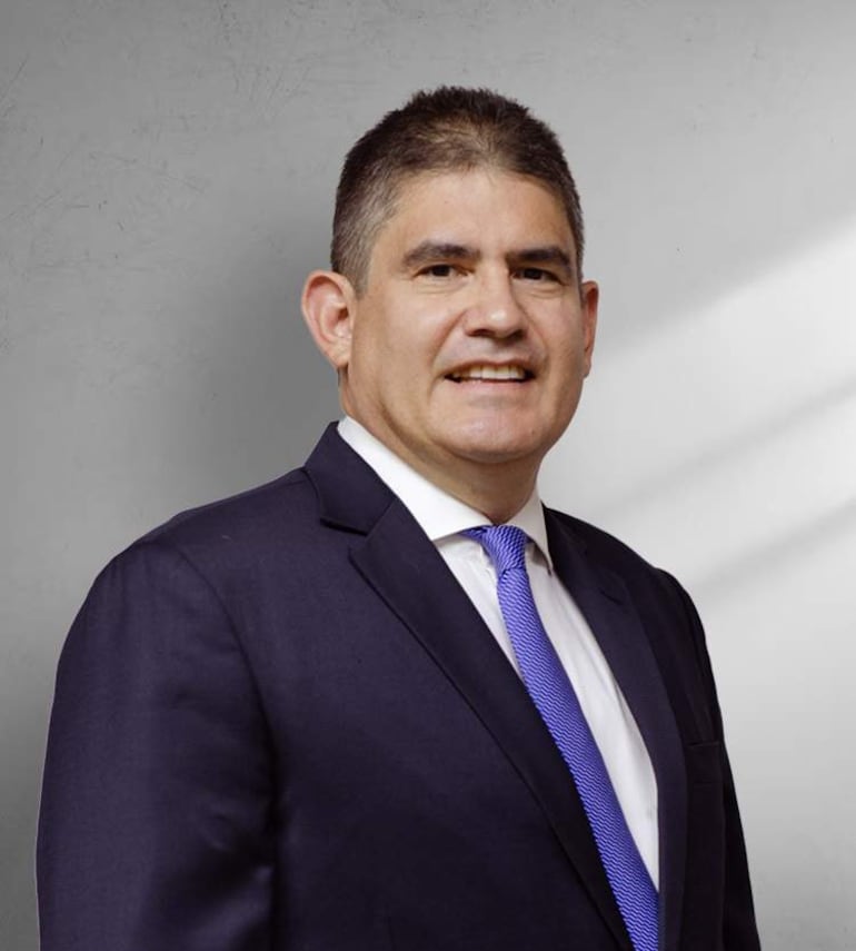 Alejandro Piera sonríe, vestido con traje oscuro y corbata azul, frente a un fondo neutro.