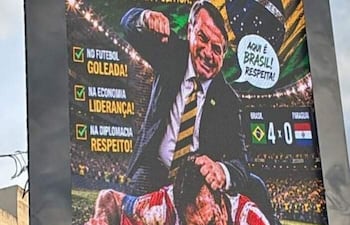 Jair Bolsonaro de pie en traje formal celebra mientras otro arrodillado con camiseta paraguaya escucha el mensaje en un cartel sobre fútbol.