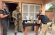 Hombre con chaleco negro escribe en cuaderno, mientras otros tres agentes, uno armado, están presentes en operativo frente a una casa.