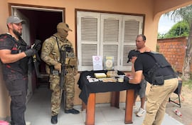 Hombre con chaleco negro escribe en cuaderno, mientras otros tres agentes, uno armado, están presentes en operativo frente a una casa.