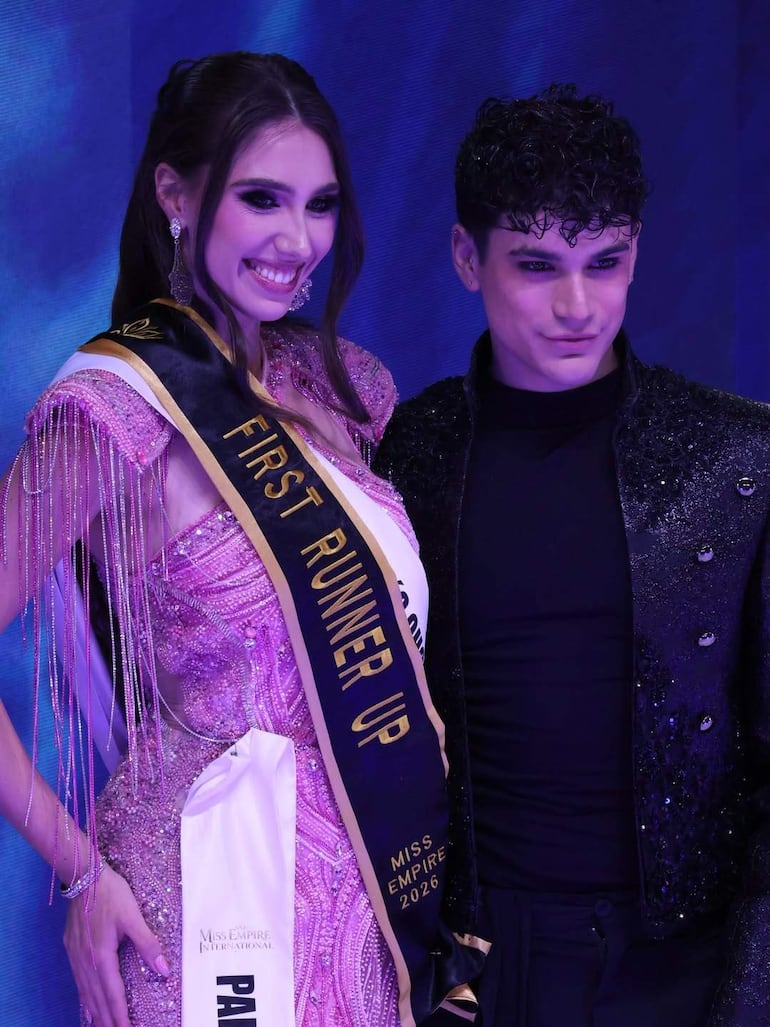 Mujer en vestido rosa brillante sonriente con banda 'FIRST RUNNER UP', junto a hombre con atuendo oscuro en ambiente festivo iluminado en azul.