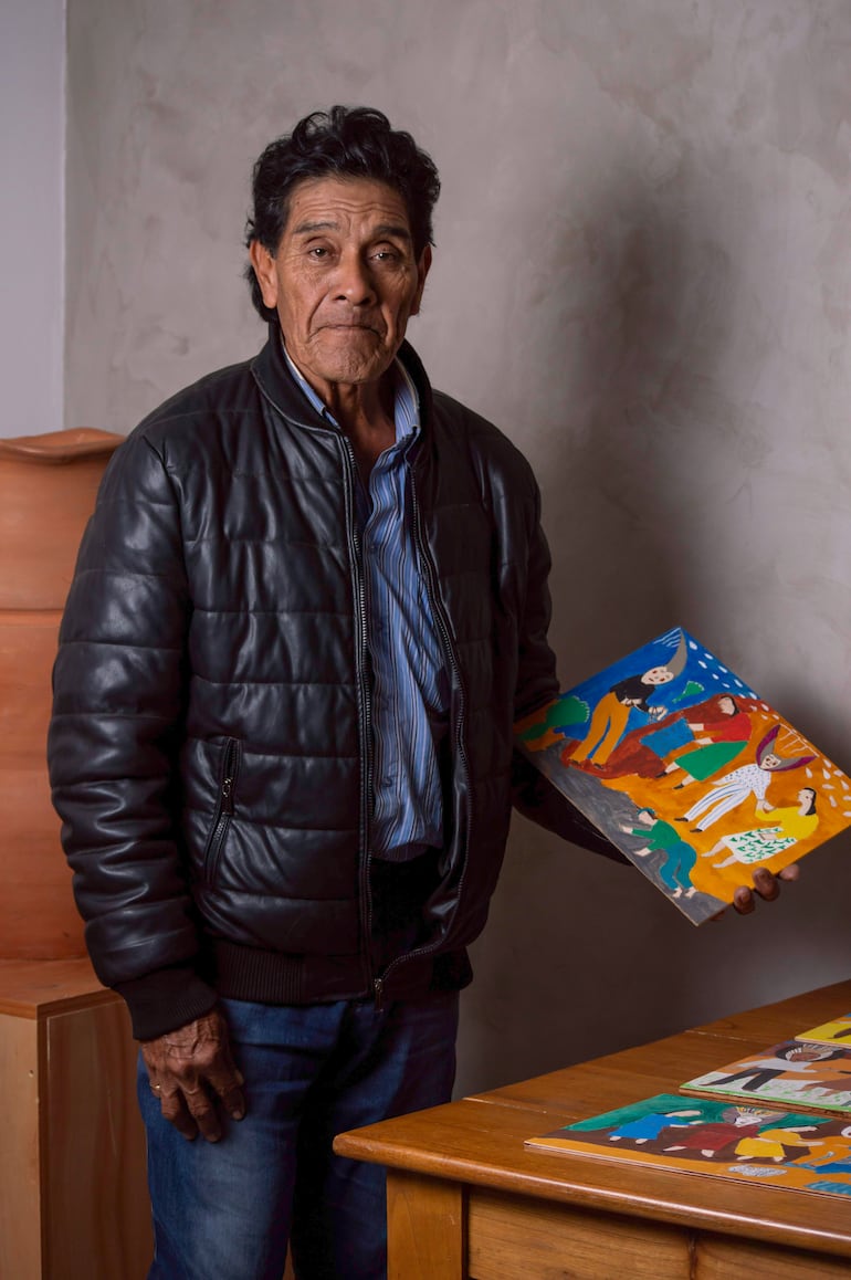 Osvaldo Pitoé de pie en chaqueta de cuero negra, sosteniendo una pintura, con un fondo sencillo y acogedor.