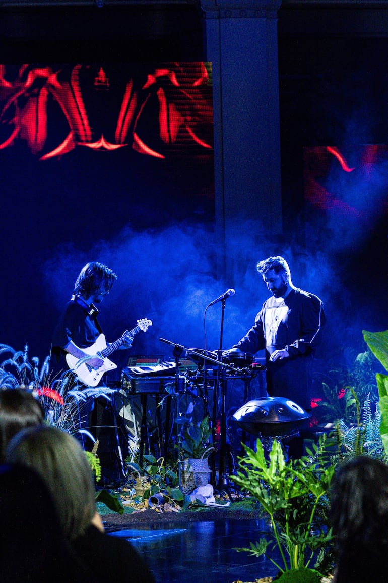 Guitarrista con cabello largo toca guitarra eléctrica blanca, junto a músico de barba en atuendo oscuro, rodeados de humo y luces de colores.