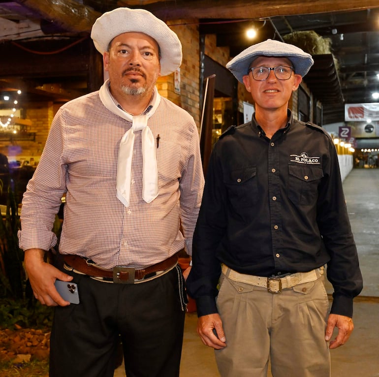 Hombres con sombreros de ala ancha, una camisa de cuadros y otra negra, en un ambiente de restaurante, sosteniendo un teléfono y mirando a la cámara.