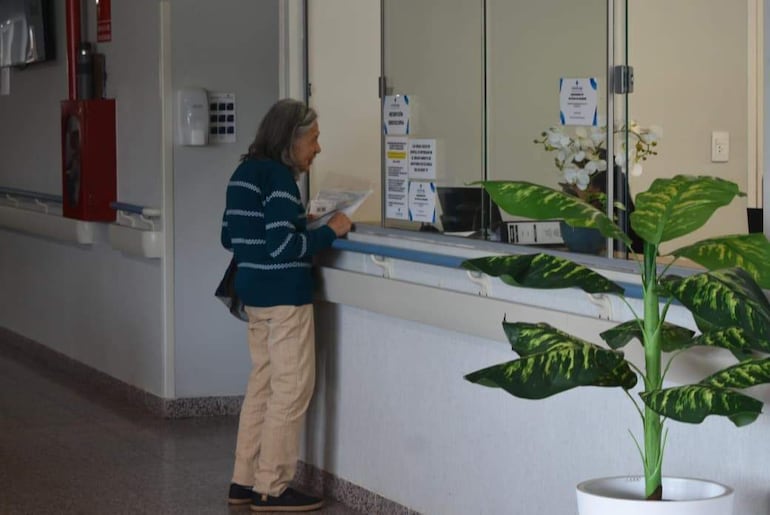 Mujer mayor con cabello canoso y suéter a rayas sostiene documentos, observando un mostrador en el hospital con decoración de planta.