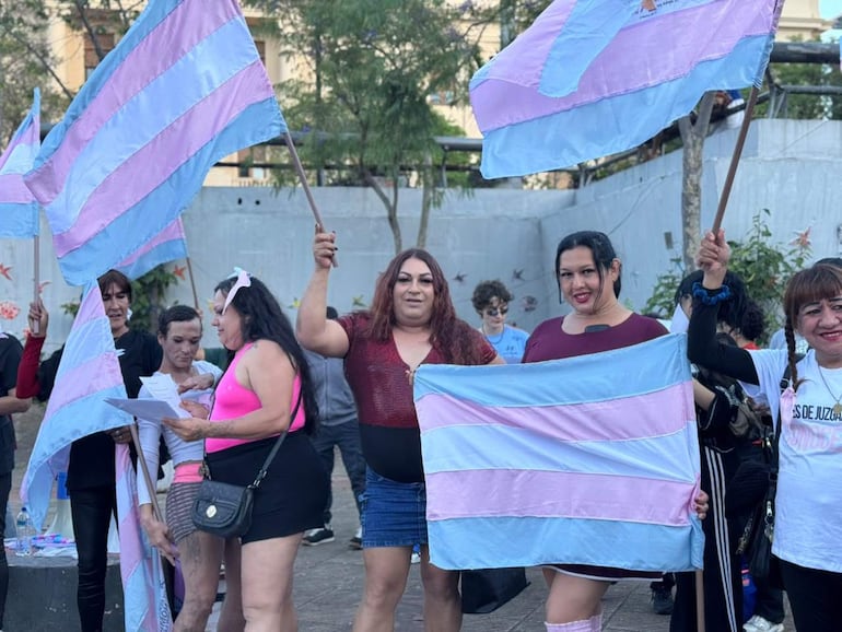 Mujeres trans con banderas azul y rosa, una sostiene un cartón, expresan alegría en la Plaza de la Democracia.